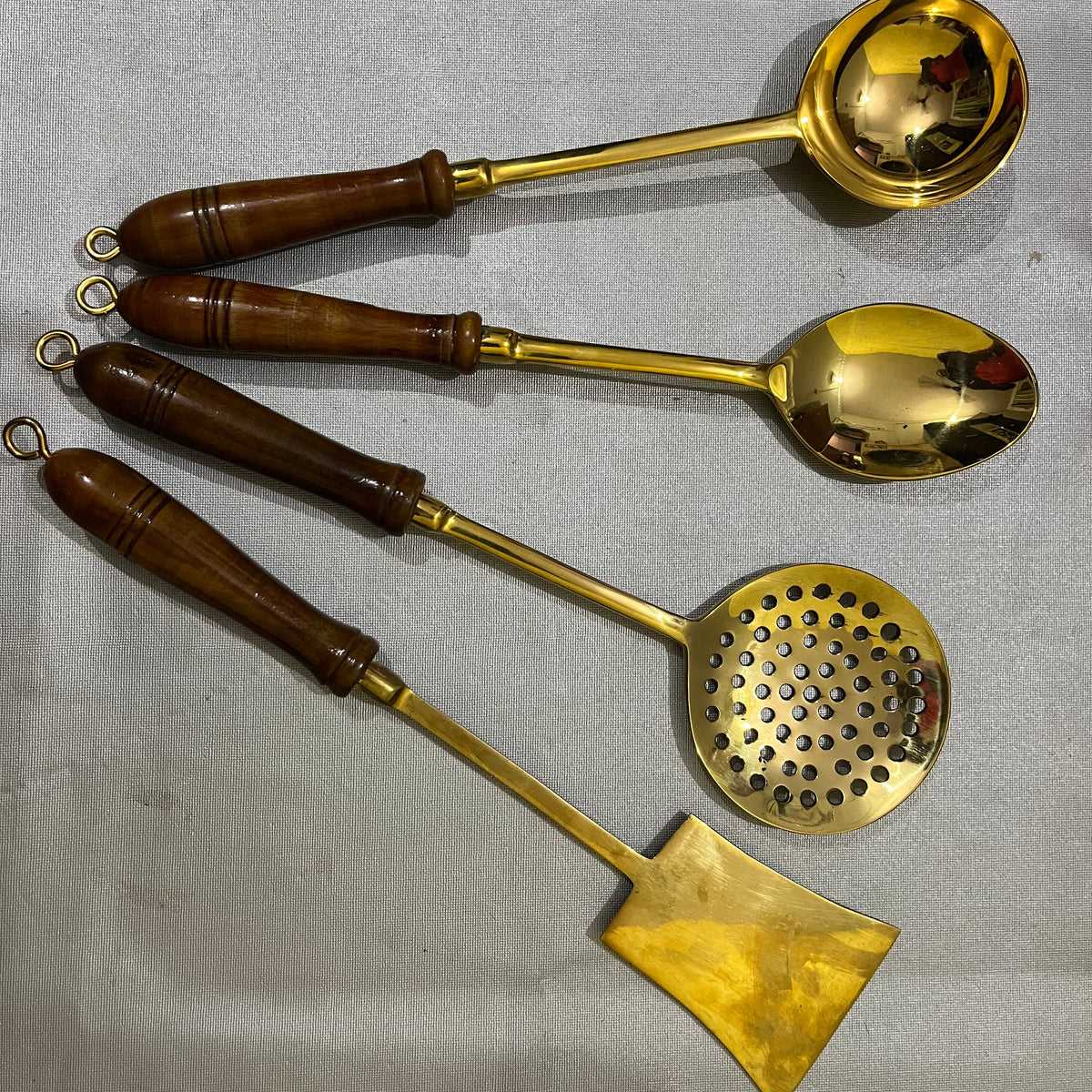 Four brass kitchen utensils with dark brown wooden handles, including a ladle, spoon, strainer, and spatula, 